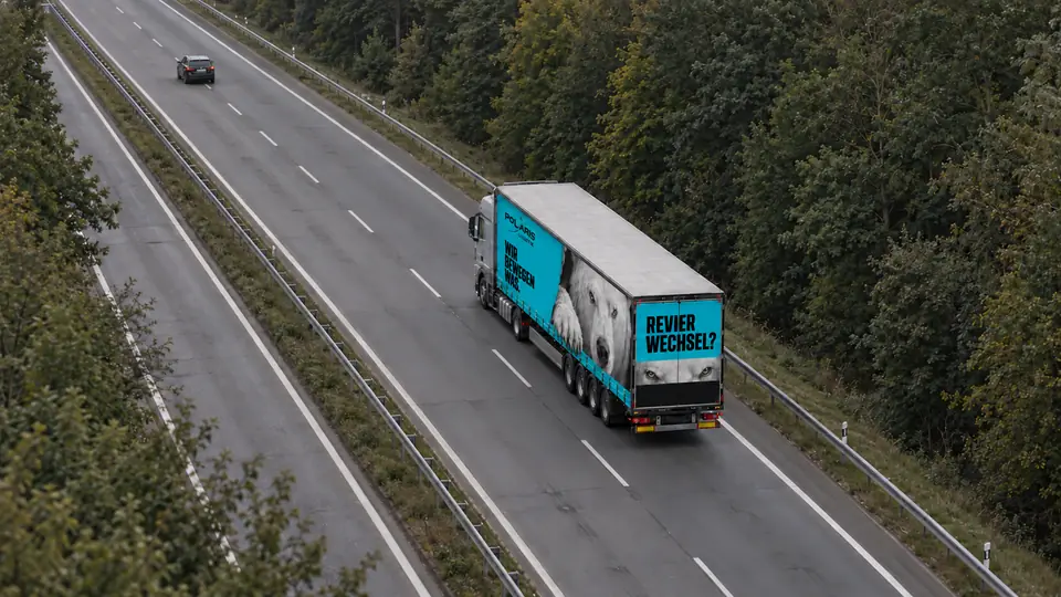 Ein Lkw mit türkisem Auflieger fährt von oben gesehen auf einer Straße durch den Wald