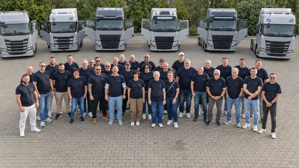 Eine große Gruppe in dunklen T-Shirts steht vor einer Reihe von Lkw auf dem Betriebshof